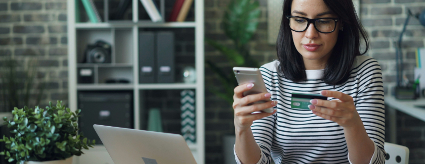 Woman sitting before her open laptop, holding her phone in one hand and credit card in another